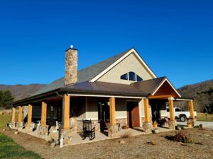 Finished SIP home with tan board-and-batten siding, stone chimney, and a wraparound porch with timber posts on stone bases, set against a mountain backdrop.